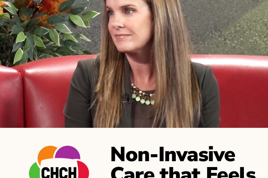 Woman with long brown hair wearing a black jacket and necklace, seated on a red couch during a discussion about non-invasive care.