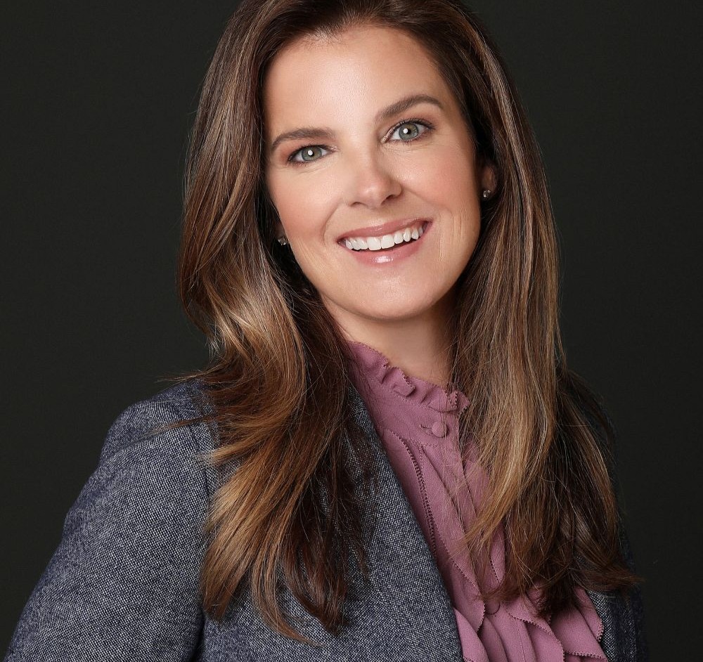 Woman with long brown hair and a warm smile, wearing a gray blazer over a purple blouse, posed against a dark background.